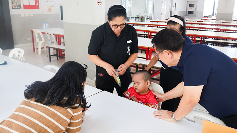Picture 4: Staff members from the Bangkok School for the Blind (left) and Mr. Piroon Laismit, Executive Director of APCD (right) interact closely with a young student in a dining hall, with Ms. Wilailekha Somprach lean in to talk with the child seated across the table.